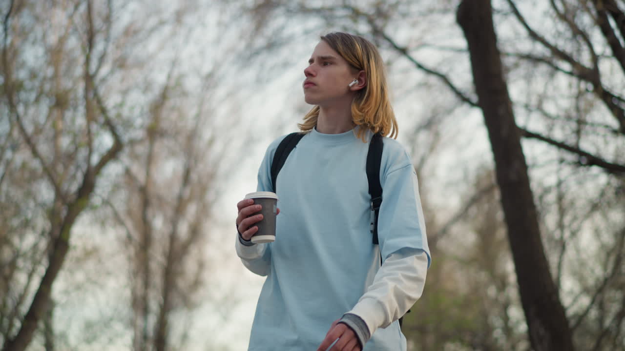 공원에 있는 학생, 공원 장면을 거니는 평범한 학생, 커피와 백팩을 들고 조용히 아침 산책을 하는 학생, 음료를 들고 공원을 거닐며 평화로운 아침 풍경을 연출하는 학생