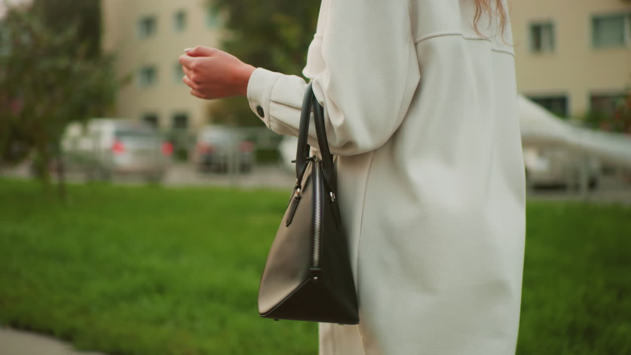 side view of woman in white coat holding black handbag walking along paved path while twirling umbrella playfully during sunset with blurred green grass, trees and residential buildings in background