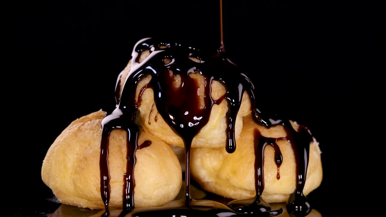 Rich chocolate sauce cascades over stacked cream-filled choux pastries on a glossy black background, captured in slow motion with dramatic studio lighting