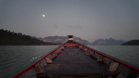 A-boat-gliding-across-the-waters-of-Khao-Sok-National-Park-in-Surat-Thani,-Thailand,-during-the-evening,-with-the-moon-shining-visibly-on-the-horizon,-embodying-the-concept-of-travel-and-vacation