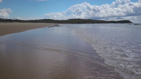 Drone-flying-over-shoreline-of-Emerald-beach-with-Look-at-me-now-Headland-in-background,-Australia