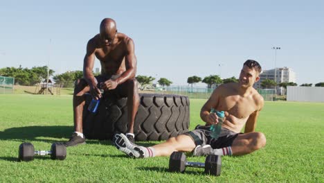 Two-fit,-shirtless-diverse-men-resting,-drinking-water-and-bumping-fists-after-exercising-outdoors