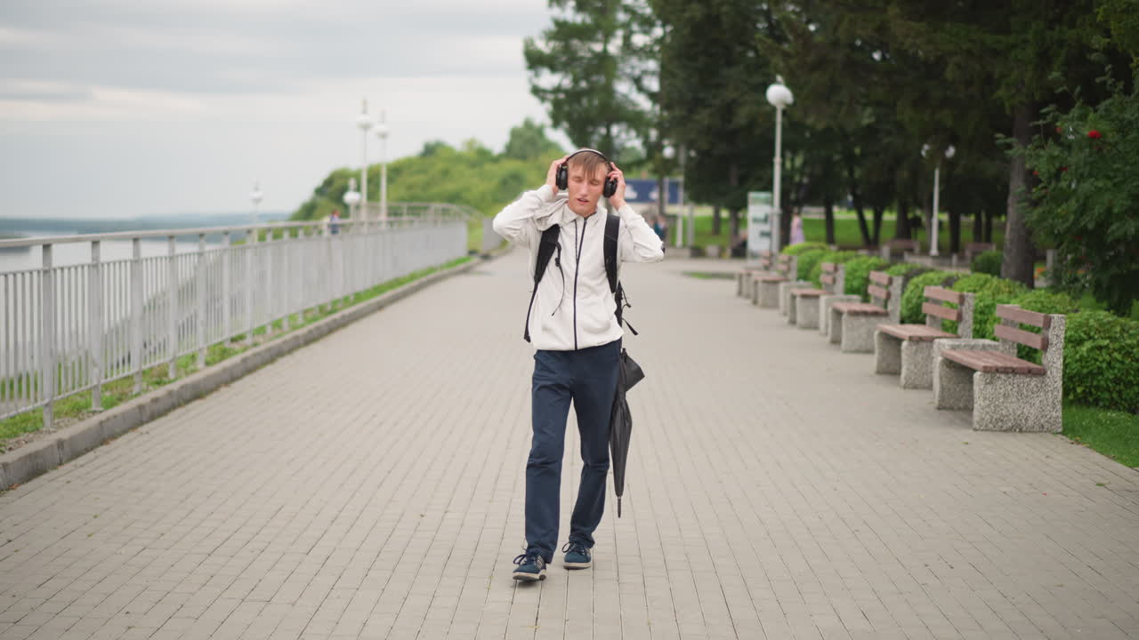 Man walking by water edge, Male traveler carries backpack along peaceful waterfront pathway under overcast sky, Man adjusting his umbrella and glasses while strolling by tranquil ocean shoreline