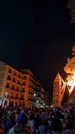 huge flames and smoke rising from a burning structure during a vibrant night festival, illuminating the faces of the captivated crowd gathered in the city square