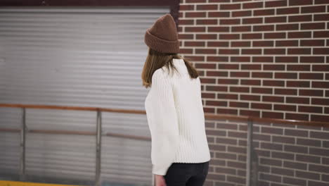 una vista de cerca de una mujer patinando tranquilamente en una pista de hielo, ella está usando un suéter blanco y un gorro marrón, con una pared de ladrillo y algunos globos coloridos