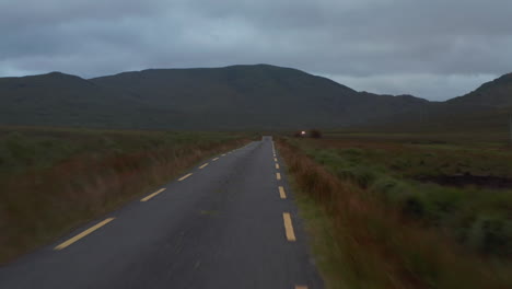 Vuelo-Bajo-Sobre-Camino-Angosto-Con-Líneas-Discontinuas-En-El-Costado.-Tiro-De-Paisaje-Con-Pastizales-Y-Montañas-En-El-Fondo.-Irlanda