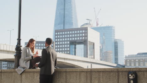 Two-millennial-colleagues-take-a-break-on-the-embankment-eating-and-talking-near-London-Bridge-by-the-River-Thames,-low-angle