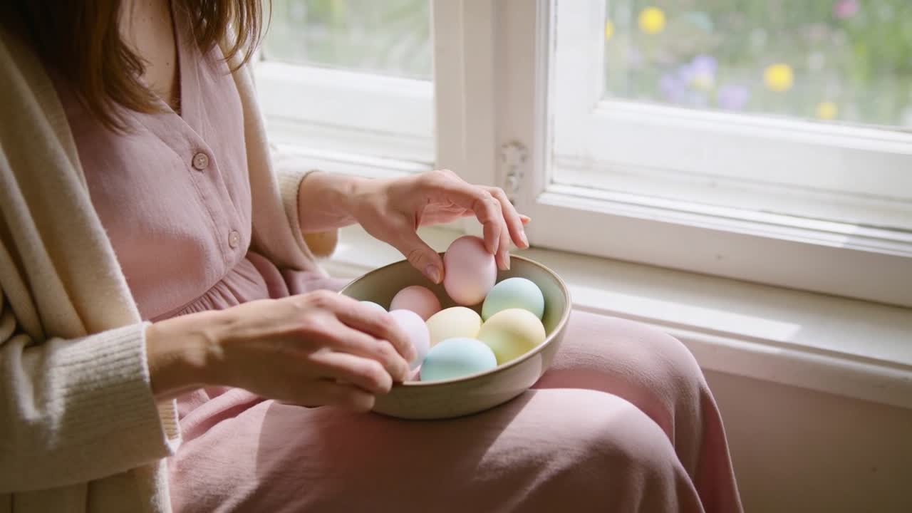 Woman placing bowl on lap by window lifting and inspecting pastel eggs tidying for home decor