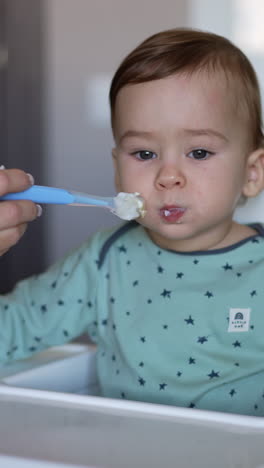 caring mother is feeding her baby boy from a spoon. mom's hand giving a spoon to her kid sitting in a feeding chair. blurred backdrop. vertical video