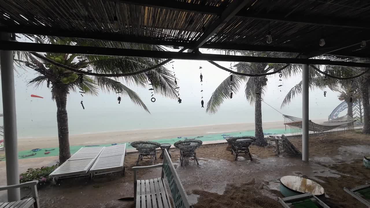 Gentle tropical rain falls over a hammock between palm trees at a quiet seaside beach, creating a serene tropical retreat with wet sand, rattan chairs, and calm ocean views