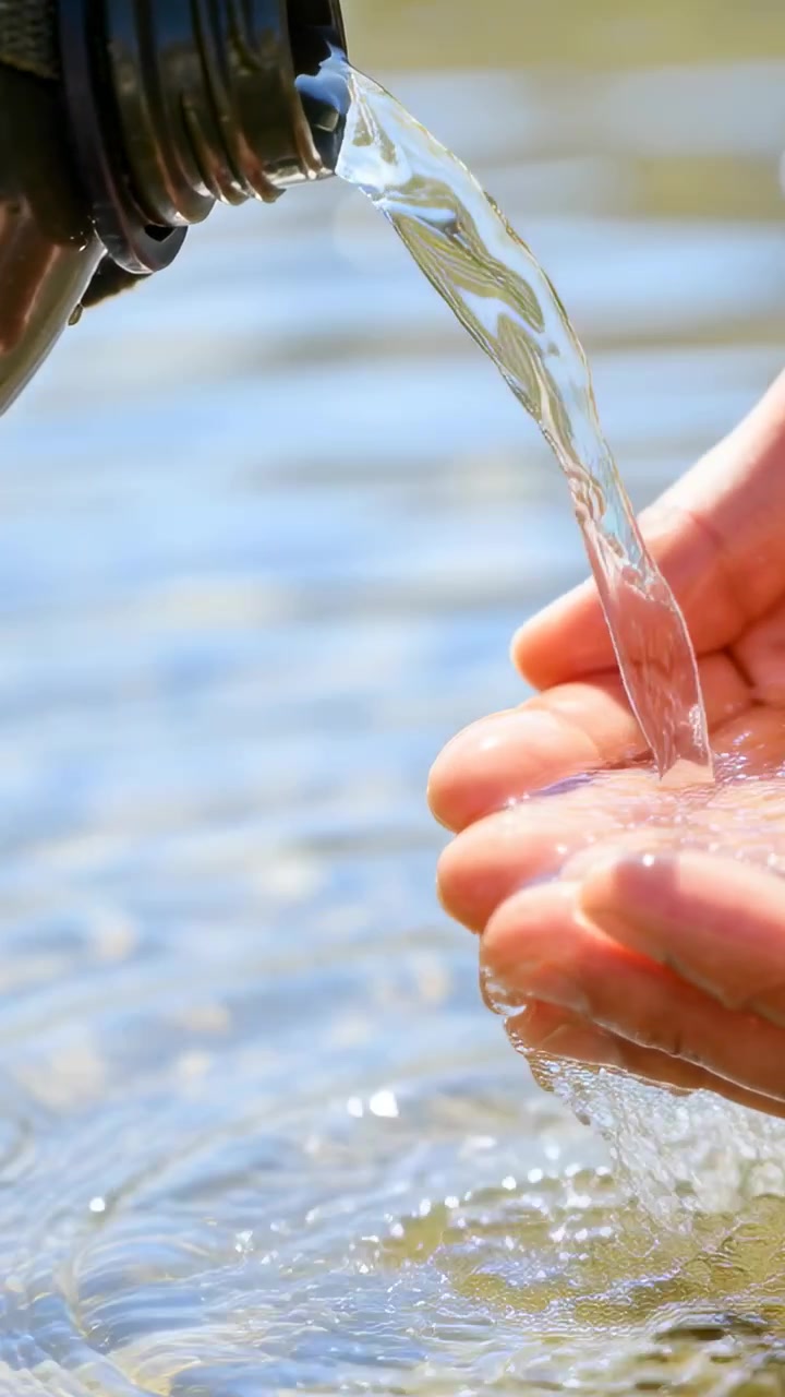 Video vertical: Inclinando una botella oscura, una mano ahuecada recogiendo agua en la orilla del lago, produciendo ondas