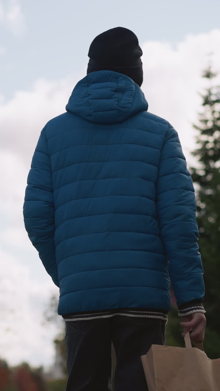 Back view of person in blue jacket and black beanie strolling in residential area holding brown paper bag, surrounded by colorful houses, trees, and bright cloudy skies on a tranquil day