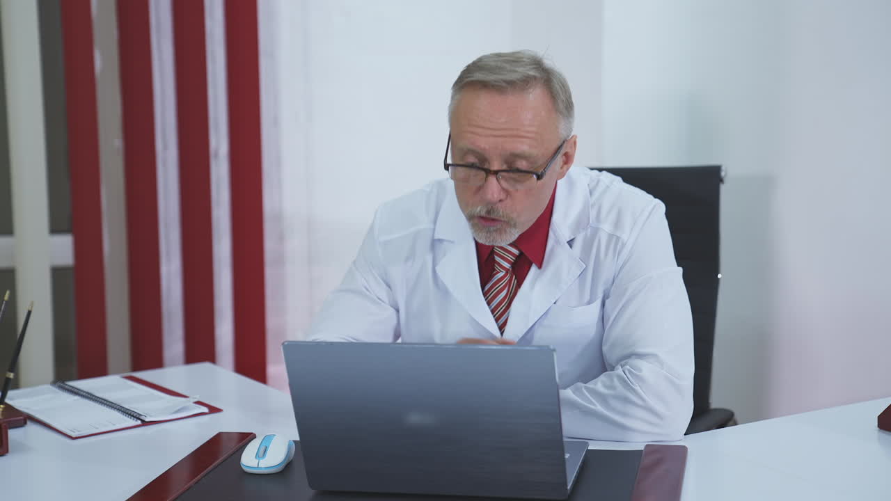 Professional doctor talks to distant patient. Male physician in white medical uniform speaking through video chat while looking at laptop in clinic.