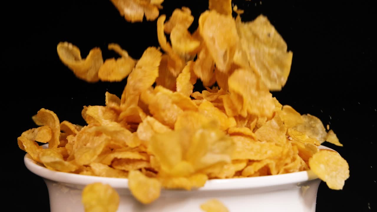 Corn flakes cascade into a white bowl against a black backdrop, captured in slow motion with dramatic lighting