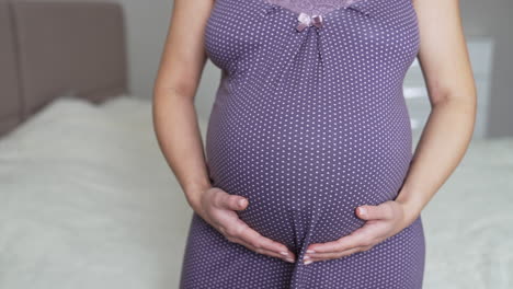adult expectant mother stands right in front of the camera. future mom strokes her big belly with two hands. her hands make a heart sign on the tummy.