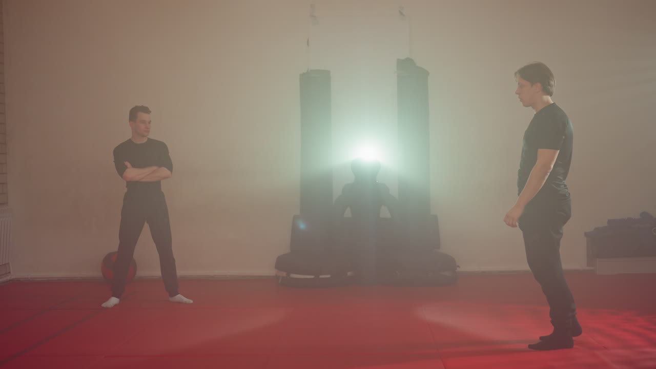 Karate practitioner performing high kick during martial arts training in gym while partner observes with arms crossed, red mats on floor, punching bags in background