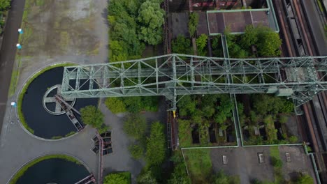 metal bridge construction spanning over grey girders that form an arch shape and are connected by smaller beams circular object which appears to be some kind of wheel or turbine trees plants around