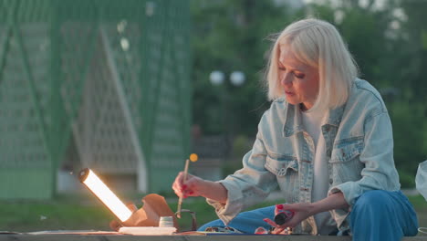 close up portrait of painter seated on bench in twilight park painting detailed strokes under lamp shine with focused expression while children play nearby framed by trees and jars of paint
