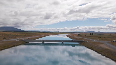 Drone-flight-over-a-blue-canal-reflecting-the-sky-while-a-car-crosses-over-a-bridge