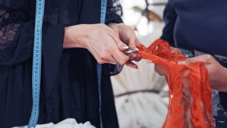 professional dressmaker is cutting red lace with scissors with the help of a tailor in atelier. woman's hands cutting out sequin and beaded red lace along its edge following the contour.