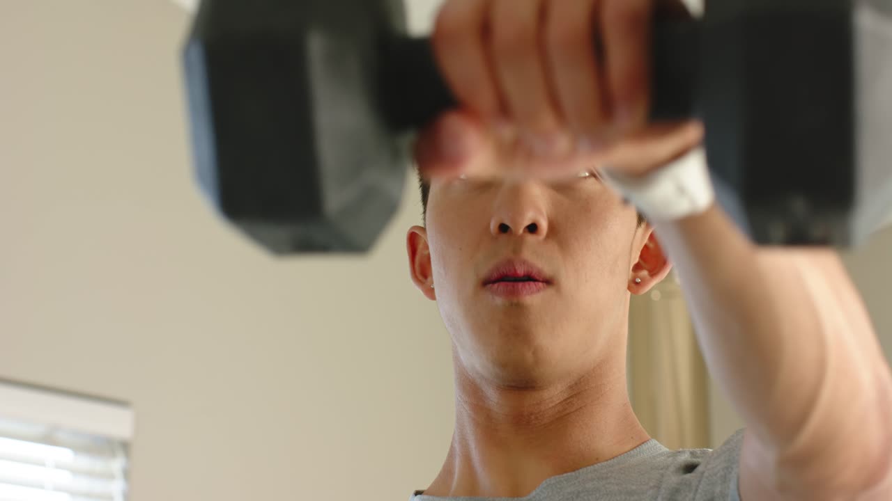 窓の近くで黒い六角形のダンベルを握り、男性が筋力トレーニングのために肩の高さまで重量を持ち上げています。