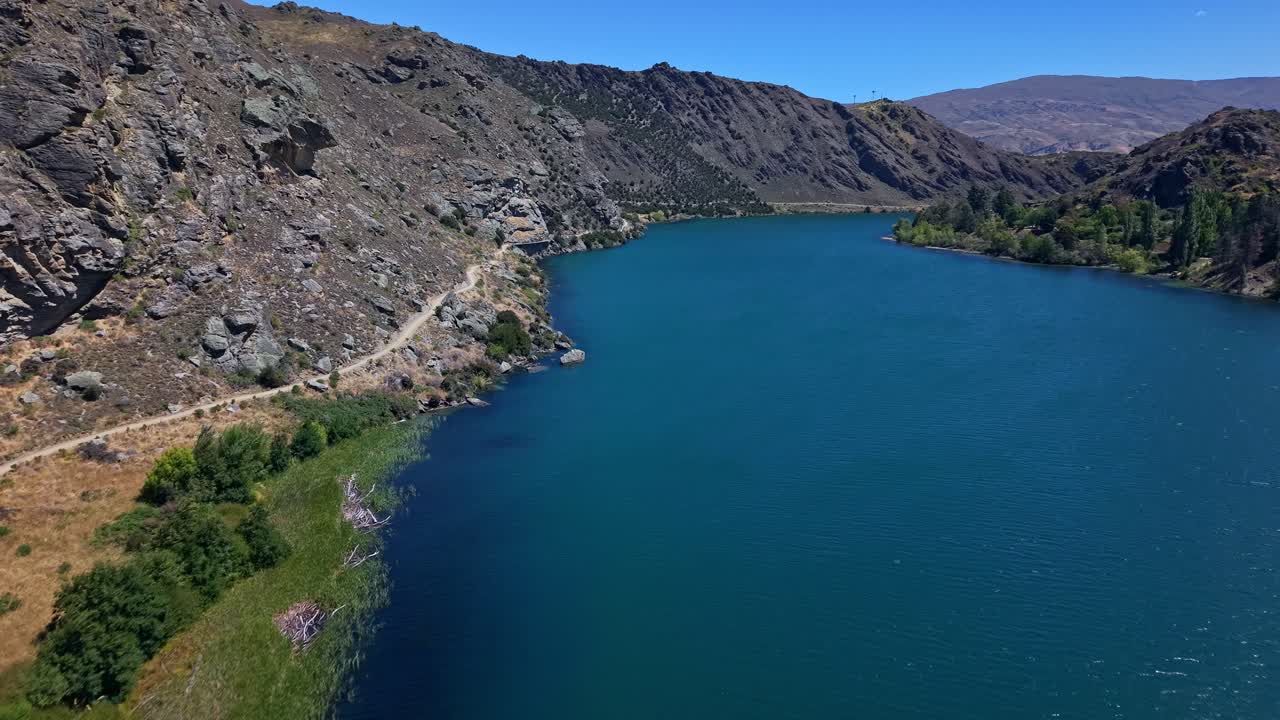 Wind blows through lush green grass and deep blue water as you travel through picture perfect new zealand gorge on beautiful summer day with this cinematic 4k drone footage of riverside cycling track