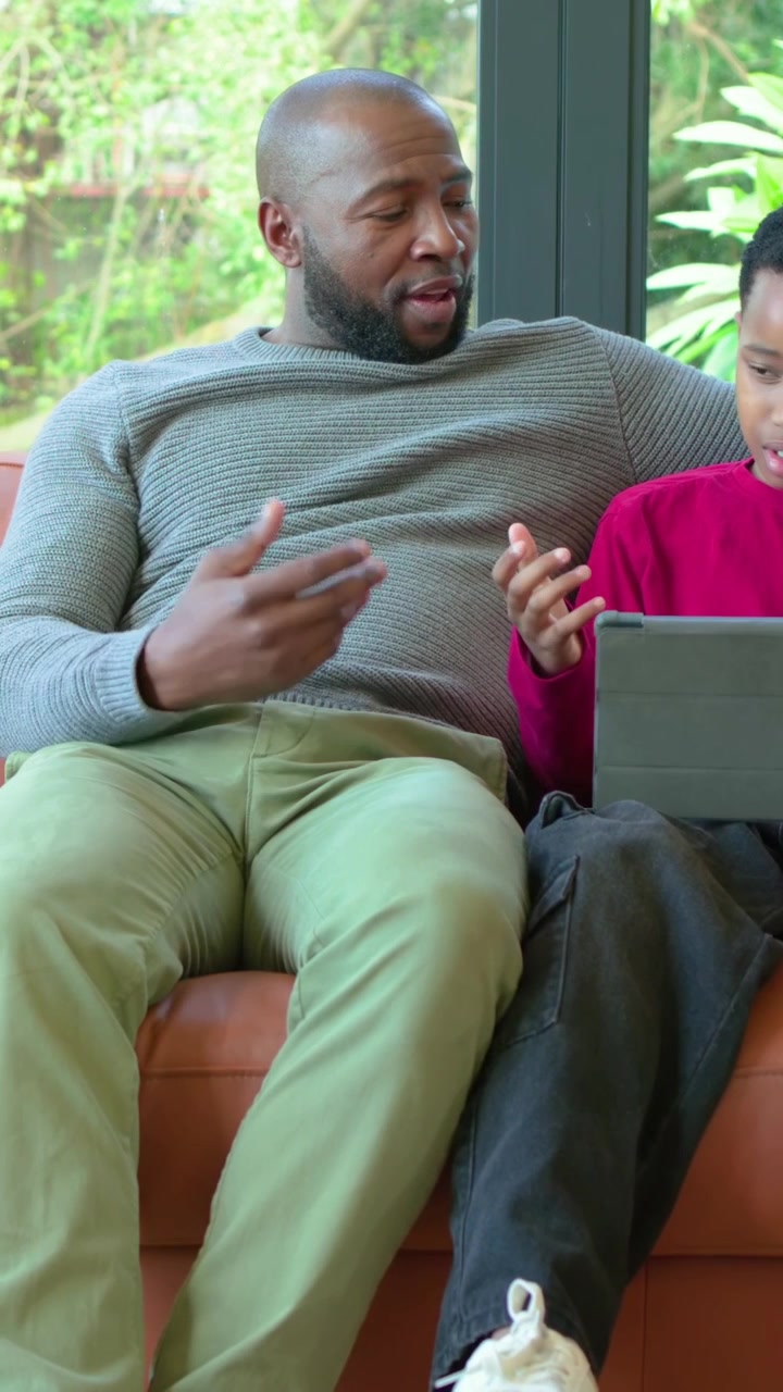 Vertical video: Reacting to tablet father leaning in guiding son on couch, pointing at tablet