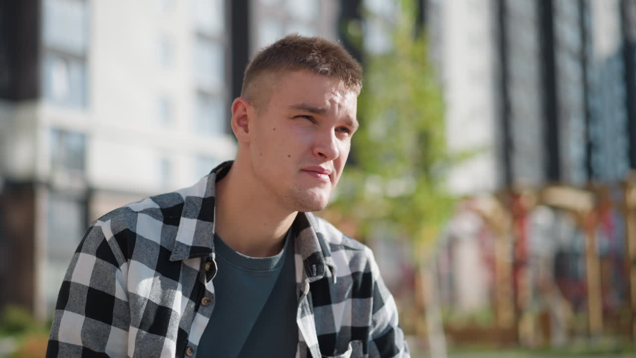 focused young student in checkered shirt places medicine in mouth while seated outdoors under bright sunlight with blurred view of greenery and tall residential buildings in background