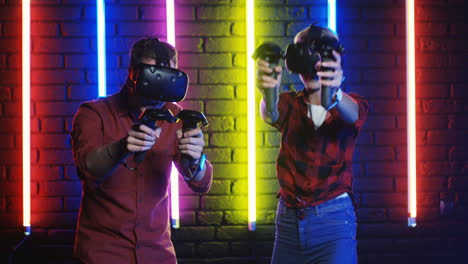 young man and woman in vr glasses and using joystick while playing a virtual reality game in a room with colorful neon lamps on the wall 1