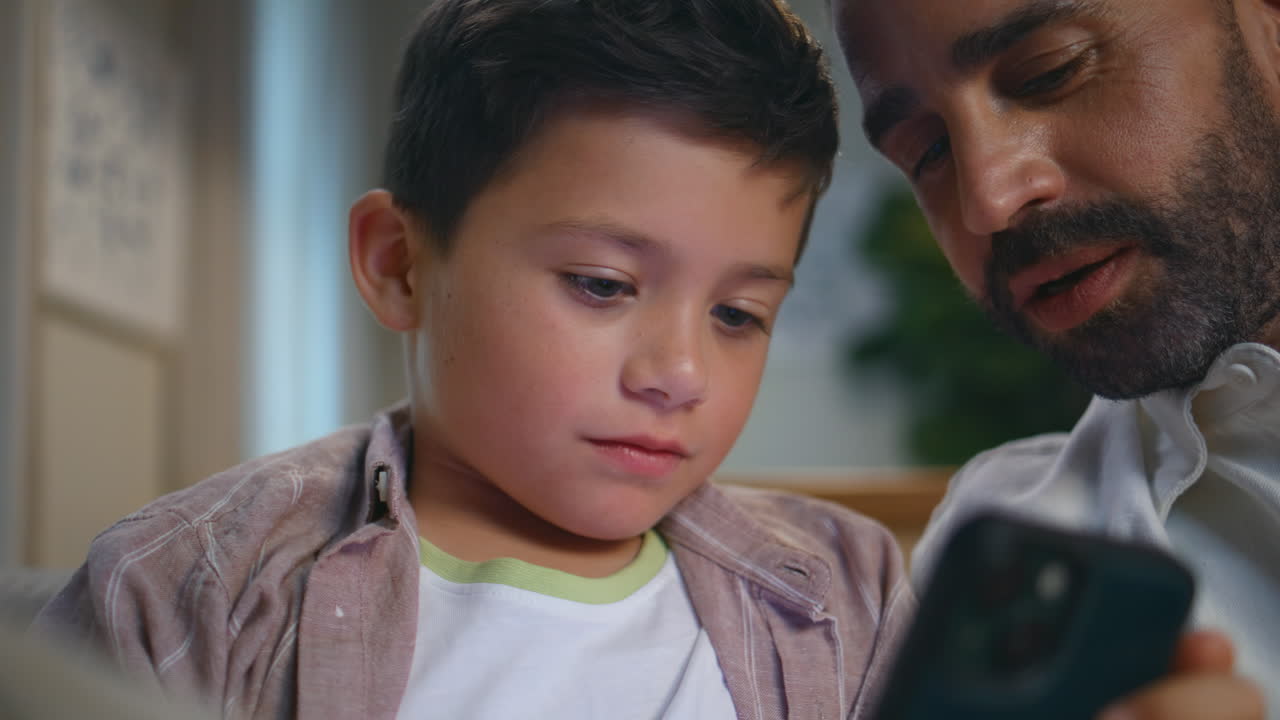 Happy boy playing mobile phone with bearded father at evening apartment closeup