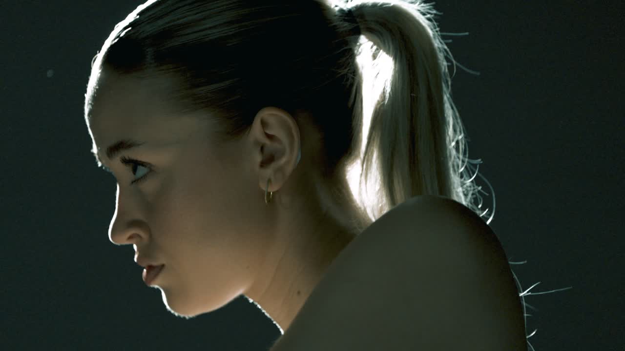 Side profile of a woman training on a stationary bike in a dark studio, with cinematic backlight wrapping her head and face in a glowing halo as she pedals with calm focus and inner strength