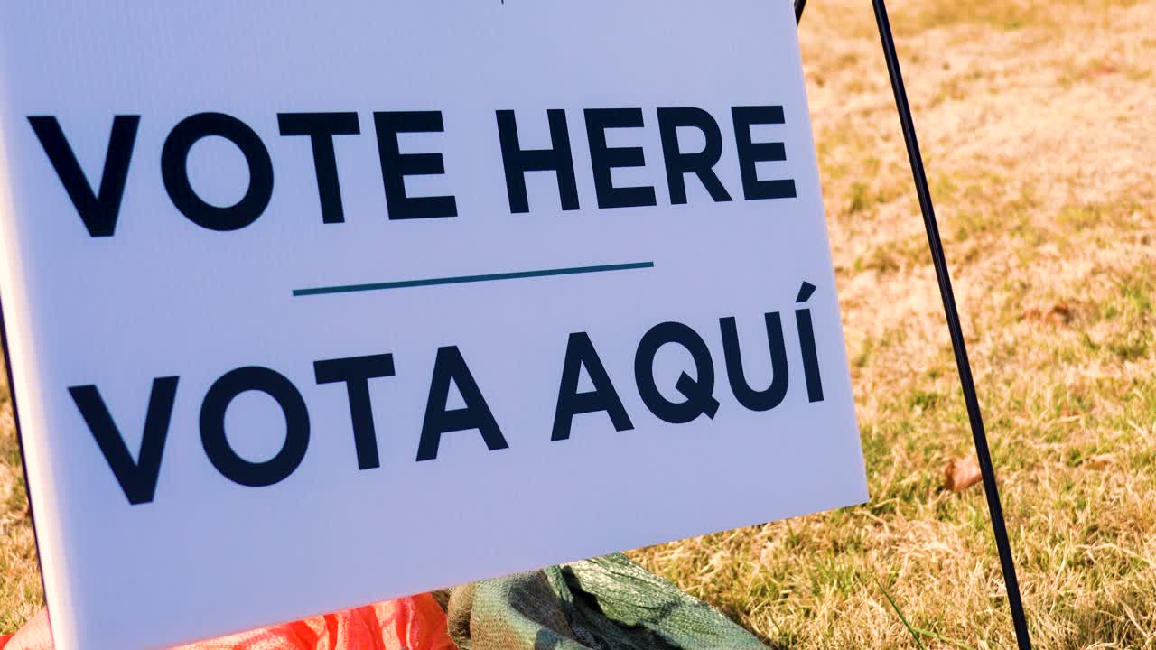 Vote Here Vota Aquí sign in close-up with bold bilingual lettering, standing out clearly as cars drive past in the blurred background. Imagery for election day, civic duty, democracy and participation