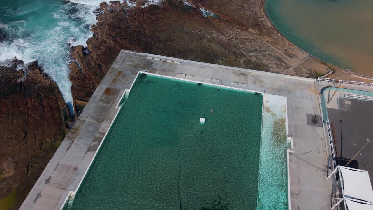 Angled aerial establishing of people enjoying Newcastle Ocean Baths with clean turquoise water and nearby beachline visible