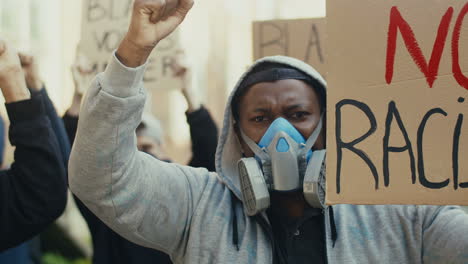 close-up view of african american man in gas mask yelling with arms up and holding a no racism" signboard in a protest with multiethnic group of people in the street"