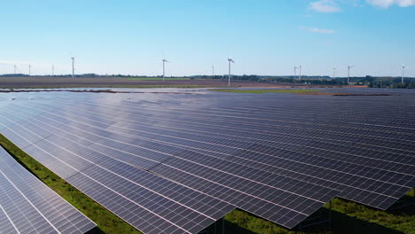 Vista-Izquierda-Del-Carro-Lento-De-Una-Granja-De-Paneles-Solares-A-Gran-Escala-Con-Molinos-De-Viento-En-El-Fondo