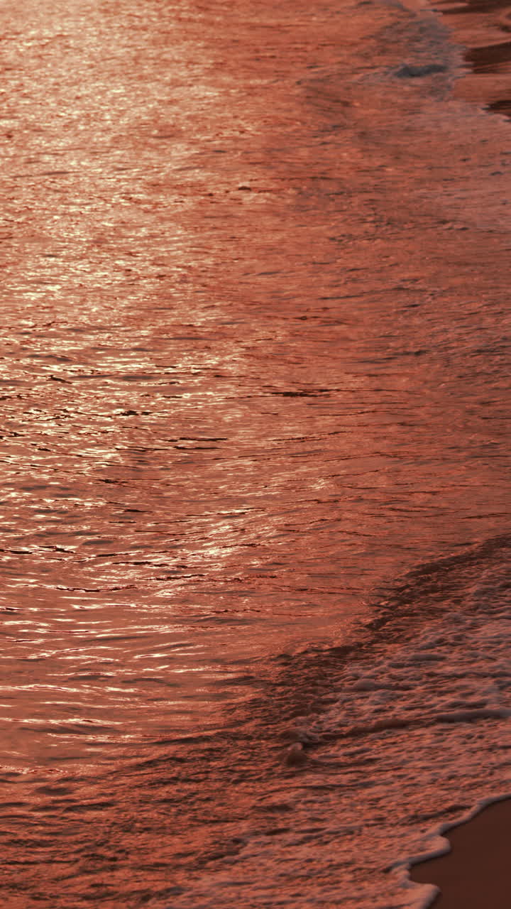Close up of gentle waves washing over the sand in golden evening light. Vertical
