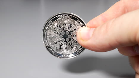 silver cardano coin held between fingers in hand on gey table