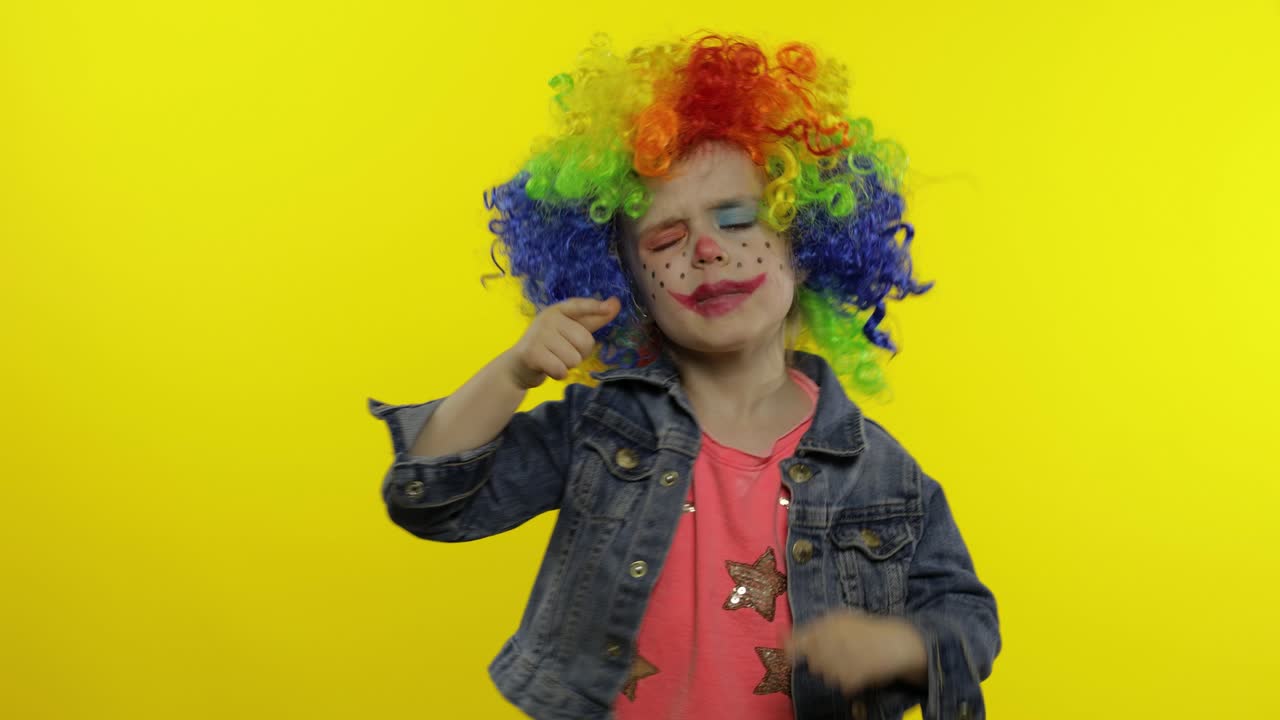 una niña payasa con una peluca arco iris haciendo caras tontas, cantando, sonriendo, bailando. halloween