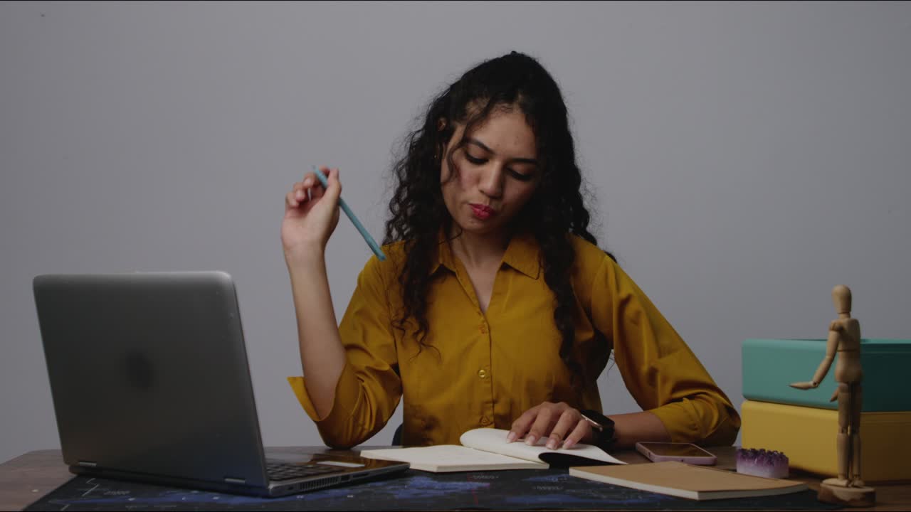 A thoughtful South Asian woman writing notes in a notebook at her office desk, pausing to think deeply during focused work