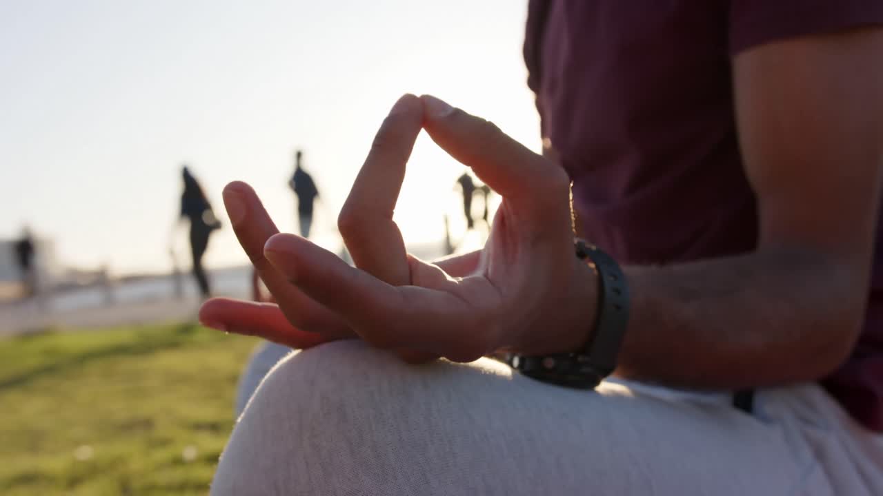 feliz hombre biracial haciendo yoga y meditando sentado en la hierba junto al mar, cámara lenta