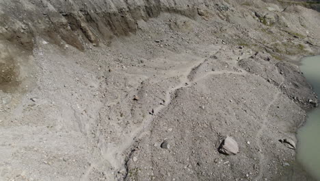 Drone-shot-of-Pasterze-Glacier-hiking-trail-and-wild-landscape,-People-walking-through-rough-and-sandy-dirt-road-alongside-the-edge-of-the-glacier-lake-at-High-Tauern-National-Park,-Austria