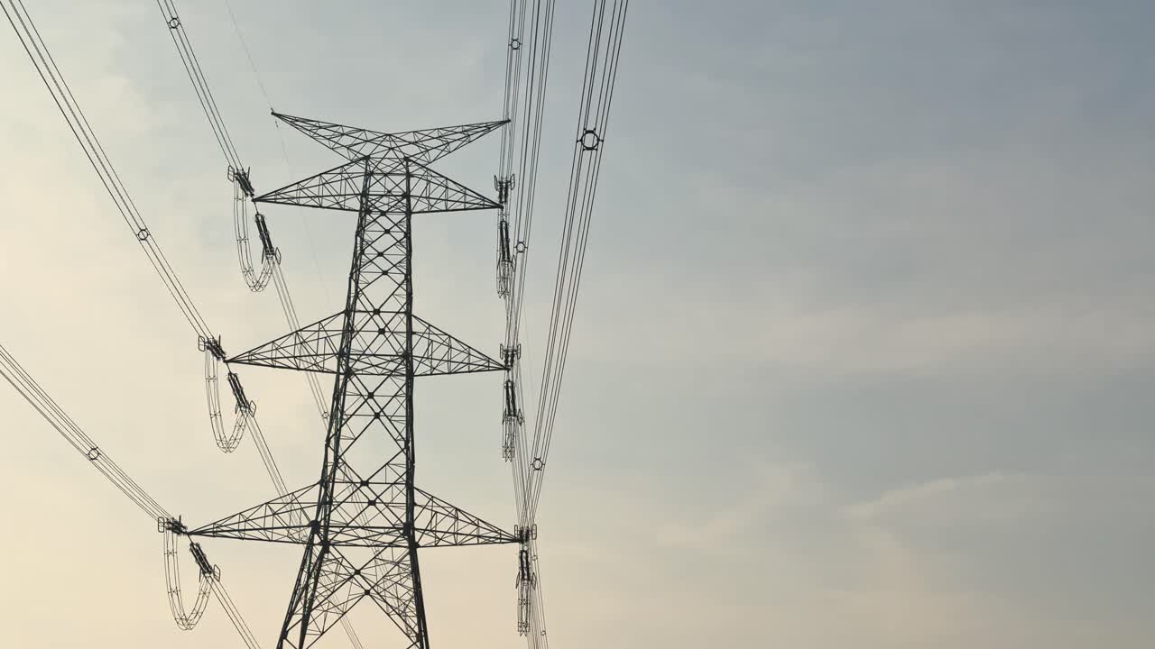 Tilt-up view reveals the full height of a high-voltage transmission tower, steel lattice rising between bundled conductors and insulators against a soft evening sky glow