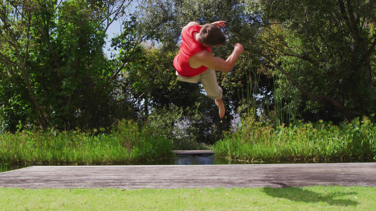 Smiling caucasian man practicing yoga, somersaulting on jetty by river with forest in background