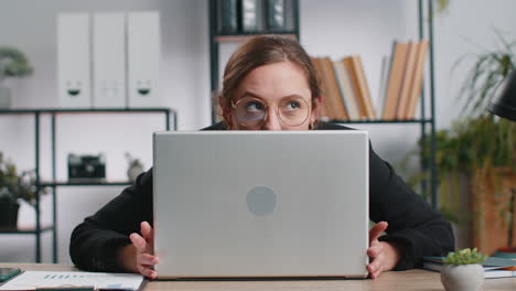 confident young business woman hiding behind laptop computer, looking at camera, spying, peeping