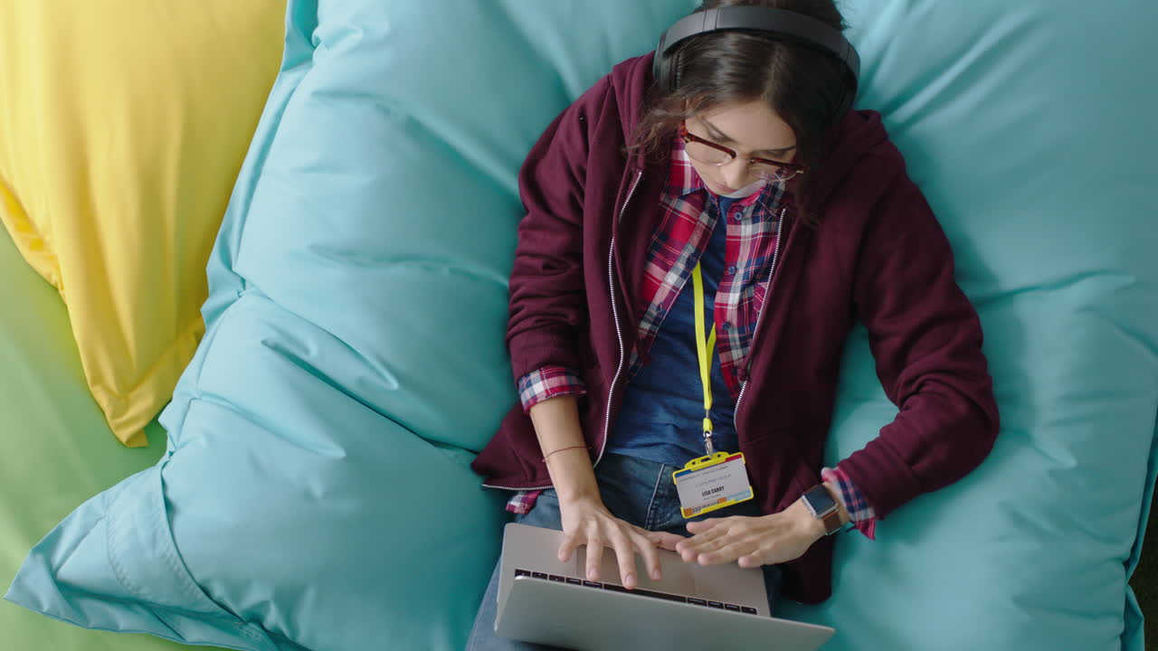 vista superior mujer feliz estudiante usando computadora portátil digital navegando en línea mensajes de redes sociales escribiendo un proyecto creativo relajándose en almohadas coloridas escuchando música en una oficina moderna