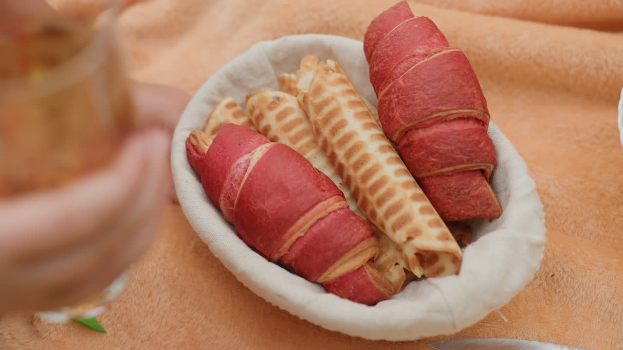 Cesta de pícnic con pastelitos enrollados, una mano cogiendo uno, vaso con bebida helada sobre una manta suave, luz dorada cálida, rollitos de barquillo crujientes y crepes de frutos rojos, ambiente de repostería casera, aperitivo relajado al aire libre
