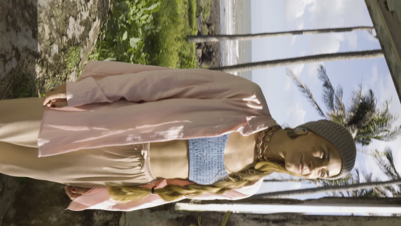 수직 비디오, 열대 낙원에서 웃고 있는 아름다운 젊은 유행 금발 모델, 화창한 푸른 하늘 날, 배경에 바다가 있는 야자수 사이에 버려진 집