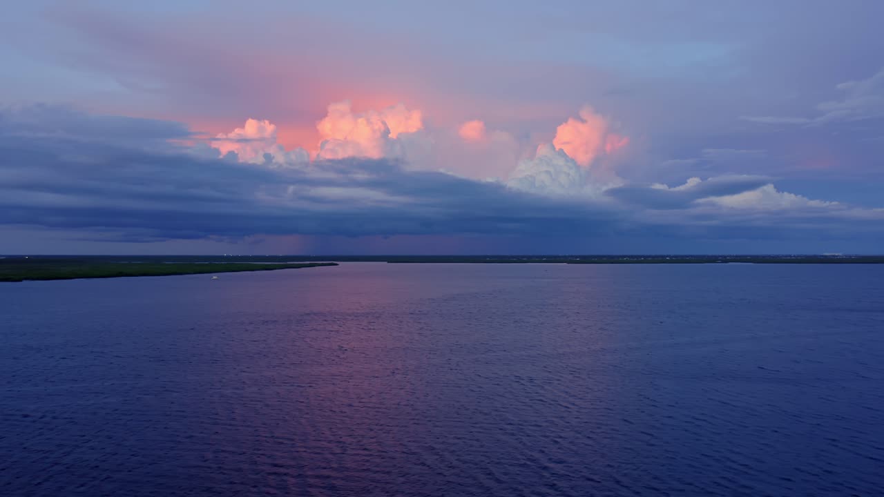 Gentle ripples move across a broad river as towering storm clouds glow pink against the twilight sky, merging calm reflection and distant energy in a serene atmospheric moment