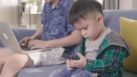 father working on laptop while child uses a smartphone on couch