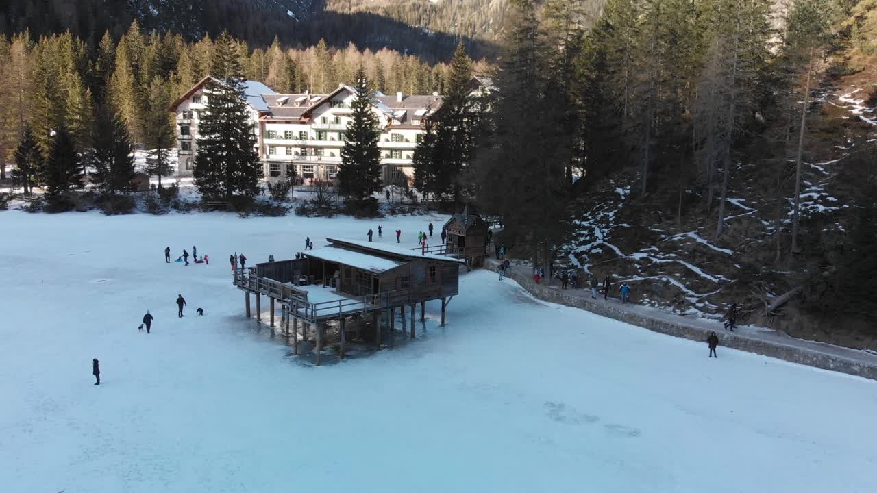 vista aérea lenta hacia abajo de la gente caminando cerca de una antigua estructura de madera en el lago braies durante el invierno en italia